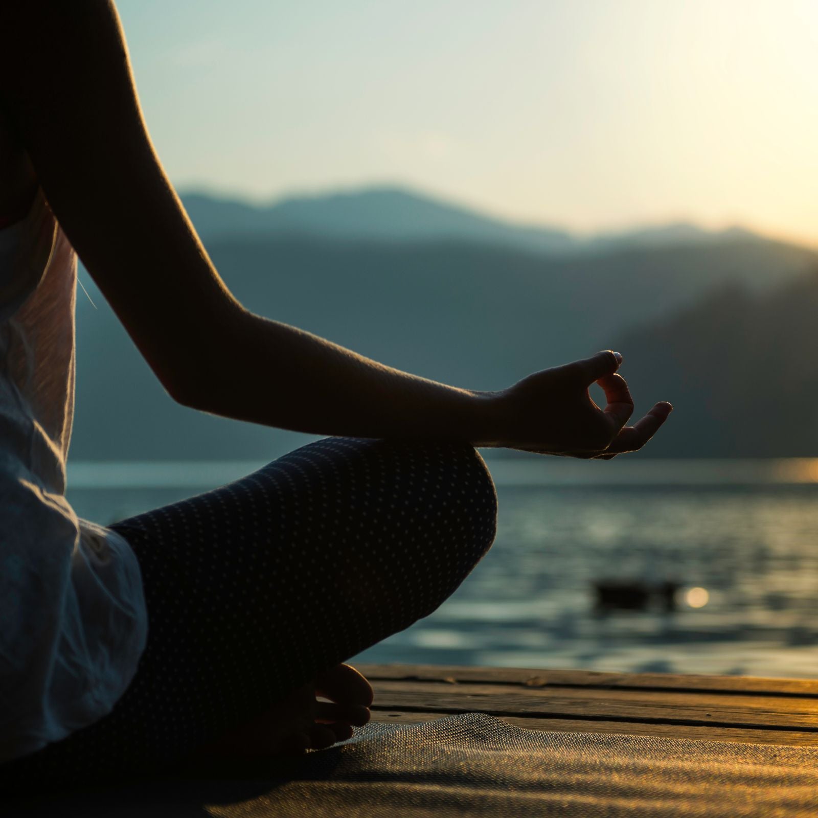 Woman meditating by a calm lake at sunrise, sitting cross-legged with hand in a relaxed mudra pose — a quiet, grounding moment in nature.